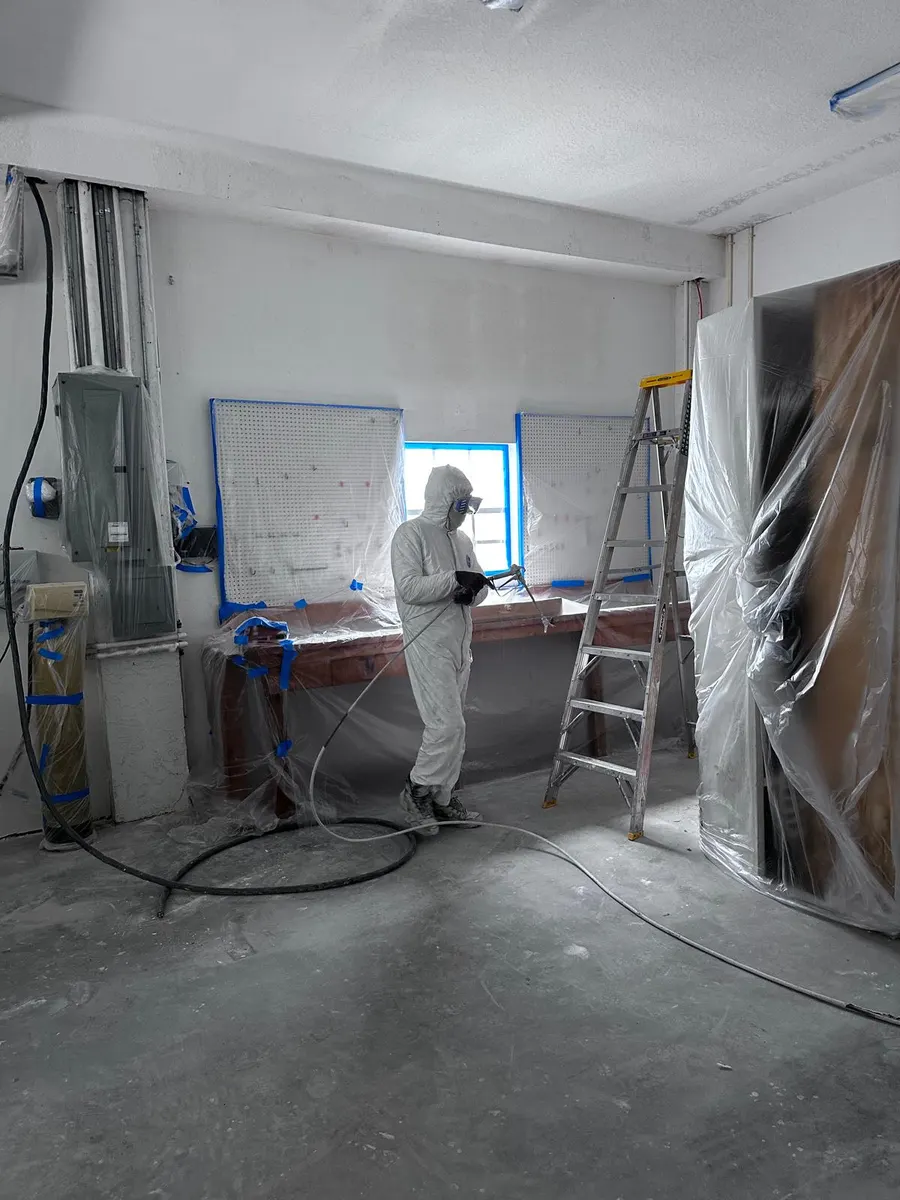 Pintor con traje de protección pintando con pistola las paredes de un espacio cubierto con plástico y cinta